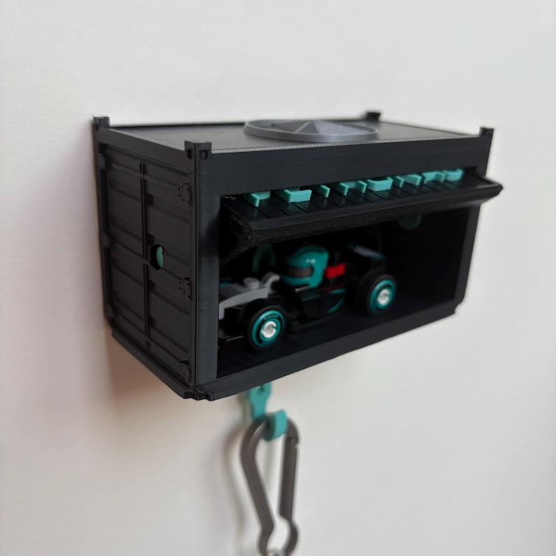 May include: A black miniature shipping container with a hinged door, housing a small toy race car. The container has teal accents and a silver carabiner clip attached. The toy car is black and teal.
