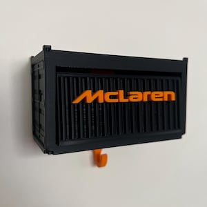 May include: Black and orange McLaren-branded wall-mounted storage container. The container has a rectangular shape with the word "McLaren" in orange across the front. A small orange hook is visible below the container.