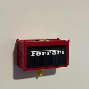 May include: Red container-shaped wall decor with the Ferrari logo in white. The container has a black panel with the Ferrari text and a yellow hook. The item is mounted on a white wall.