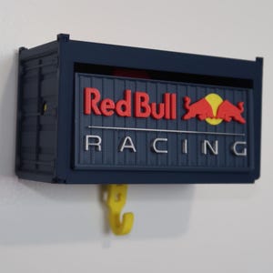 May include: A navy blue wall-mounted container with the Red Bull Racing logo in red and white. The container has a yellow hook below. The logo features the words "Red Bull Racing" and the Red Bull logo.