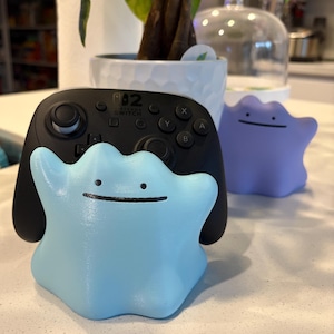 以下が含まれることがあります： 黒いNintendo Switchコントローラーが付いた、水色のDitto型コントローラーホルダー。紫色のDittoのフィギュアも表示されています。コントローラーホルダーには笑顔があります。アイテムは白い表面にあります。