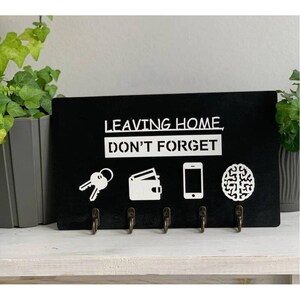 Könnte beinhalten: Schwarzer Schlüsselhalter mit dem Text "Leaving Home, Don't Forget" und fünf Haken. Der Schlüsselhalter hat Symbole für Schlüssel, eine Brieftasche, ein Telefon und ein Gehirn.