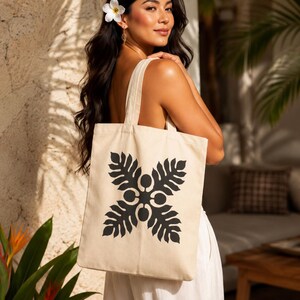 May include: A beige canvas tote bag featuring a black graphic print of four symmetrical monstera-style leaves arranged in a cross pattern. The bag has two shoulder straps and a rectangular shape, constructed from a light-colored woven fabric material.