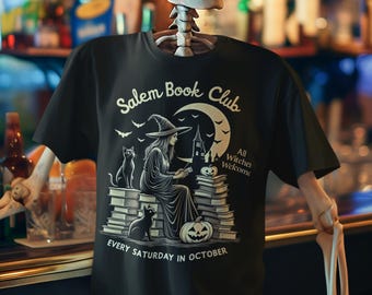 Puede incluir: Camiseta negra con una bruja leyendo sobre una pila de libros, dos gatos negros, una calabaza y una luna creciente. El texto dice "Salem Book Club", "All Witches Welcome" y "Every Saturday in October".