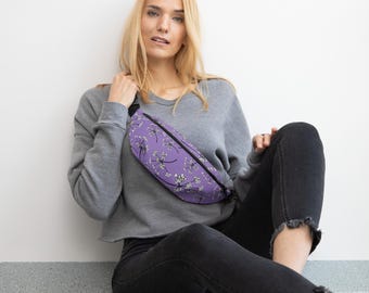 May include: A purple fanny pack with a black zipper and strap. The bag features a repeating pattern of white and black floral designs. The fanny pack is worn across the body.