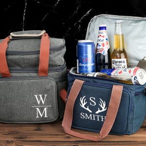 Peut inclure: Deux sacs isothermes personnalisés. L'un est gris avec les initiales "W/M" et l'autre est bleu avec le texte "S SMITH" et des bois de cerf. Les deux sacs ont des sangles brunes et sont remplis de boissons.