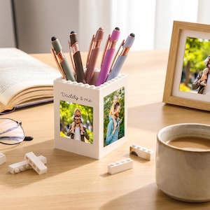 Può includere: Un portapenne bianco con la scritta "Daddy & me" e una foto di un padre e un bambino. Il portapenne contiene diverse penne con dettagli oro rosa. Una foto incorniciata, una tazza di caffè e un libro aperto sono anche sulla superficie di legno.