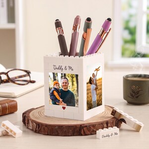 Op de afbeelding: Een witte pennenhouder met foto's en de tekst "Daddy & Me" op een houten voet. De pennenhouder bevat verschillende pennen in bruin-, roze- en paarse tinten. Een kopje koffie en een bril staan ook op tafel.