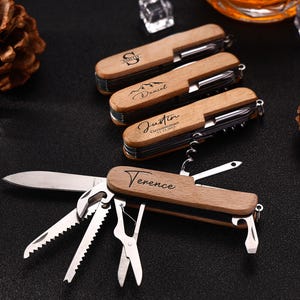 Puede incluir: Multiherramientas de madera con grabados personalizados. Las herramientas tienen una variedad de funciones, incluyendo un cuchillo, sierra y sacacorchos. Los nombres "Terence", "Justin", "Daniel" y "S" están grabados en los mangos de madera. Las herramientas están sobre una superficie negra.