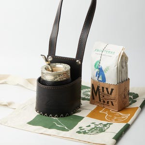 May include: A dark brown leather mate cup holder with a silver mate cup and straw. A bag of yerba mate is in a small cardboard box. The items are on a patterned cloth tote bag.