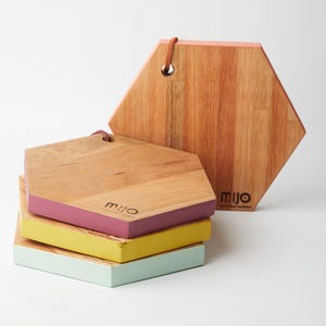May include: Four hexagonal wooden cutting boards stacked on top of each other. The top board is natural wood with a light pink edge. The next board is natural wood with a purple edge. The third board is natural wood with a yellow edge. The bottom board is natural wood with a light green edge. Each board has the word "MIJO" printed on it.
