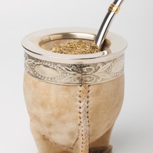 Può includere: Un contenitore per mate in pelle beige con un bordo in metallo argentato caratterizzato da un motivo floreale in rilievo. Il contenitore poggia su tre piedini rivestiti in pelle e contiene erbe verdi essiccate. È inserita una cannuccia in metallo argentato con una fascia dorata.