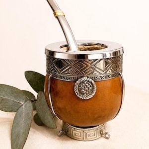 Può includere: Un contenitore per mate in zucca marrone con bordo, base e dettagli decorativi in metallo argentato. La tazza presenta un motivo geometrico sulla fascia superiore e un emblema circolare sul davanti. È inserita una cannuccia in metallo argentato con un anello dorato.
