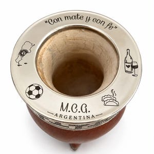 Pode incluir: Uma cuia de mate de madeira castanha com um rebordo de metal prateado gravado com o texto "Con mate y con fe" e "M.C.G. ARGENTINA". O rebordo apresenta ícones gravados de um mapa da Argentina, uma bola de futebol, uma garrafa de vinho e empanadas.