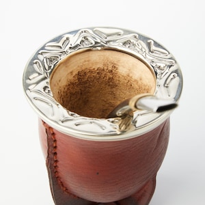 Puede incluir: Una calabaza de cuero marrón con un borde plateado y una pajita de metal. La calabaza tiene un interior de madera y está sostenida por una base de madera marrón oscuro. El borde plateado tiene un patrón decorativo.