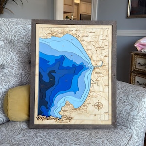 May include: A framed wooden map of Monterey Bay, California, with varying shades of blue representing the depth of the water. The map is encased in a dark brown frame and includes the text "Monterey Bay California".