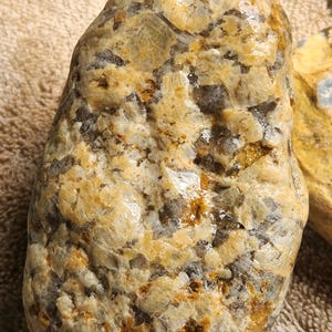 Könnte beinhalten: Ein natürlich geformter, polierter Stein mit einer Mischung aus Beige-, Gold- und Grautönen. Die Oberfläche hat ein strukturiertes Aussehen mit Bereichen kristalliner Strukturen und einem glänzenden Glanz. Die Form des Steins ist glatt und abgerundet.