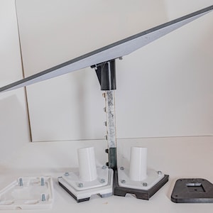 May include: A white and black solar panel mounting system with a gray panel, a black metal pole, and white plastic bases. The system is designed to be mounted on a flat surface.