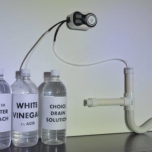 May include: A setup for a drain cleaning experiment. Three clear plastic bottles labeled "50/50 Water Bleach", "White Vinegar 5% Acid", and "Choice Drain Solution" are connected to a white PVC pipe system and a digital display.