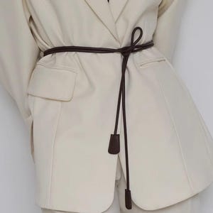 May include: Cream-colored blazer with a dark brown belt tied at the waist. The belt has a long, thin design with a small, rectangular accent at the end. The blazer has two front pockets and a classic lapel.