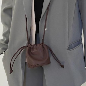 Könnte beinhalten: Ein hellgrauer Blazer mit passender Hose. Eine kleine, braune Kordelzugtasche hängt am Hals. Der Blazer hat ein klassisches Design mit fallendem Revers und einer Tasche. Das Outfit ist in einer neutralen Farbe gehalten und für verschiedene Anlässe geeignet.