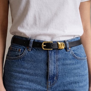 Könnte beinhalten: Ein schwarzer Ledergürtel mit einer goldfarbenen Schnalle und zwei dekorativen goldfarbenen Schlaufen. Der Gürtel wird mit einer blauen Jeans und einem weißen T-Shirt getragen. Ein klassisches Accessoire.