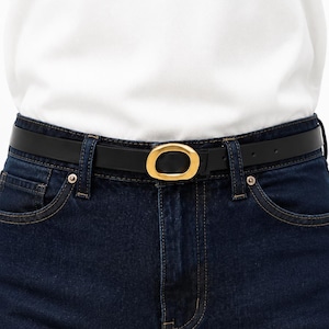 Pode incluir: Um cinto fino de couro preto com uma fivela metálica dourada de formato oval e liso. O cinto está passado pelos passadores de uma calça jeans azul-escura, servindo como um acessório limpo e funcional para prender a calça na cintura.