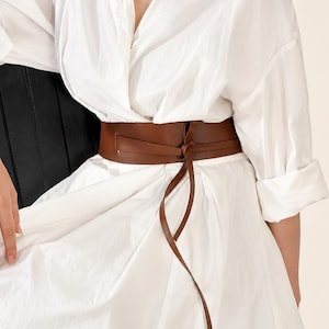 Pode incluir: Um cinto largo de couro castanho estilo obi com design envolvente e tiras longas e finas que se atam à frente, usado sobre um vestido camiseiro branco com botões, mangas com punhos e gola clássica.