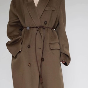 May include: An oversized olive brown wool-blend coat featuring a double-breasted front with dark brown buttons, wide lapels, and two side flap pockets. A thin, dark brown leather cord is tied around the waist as a belt, hanging down the front.