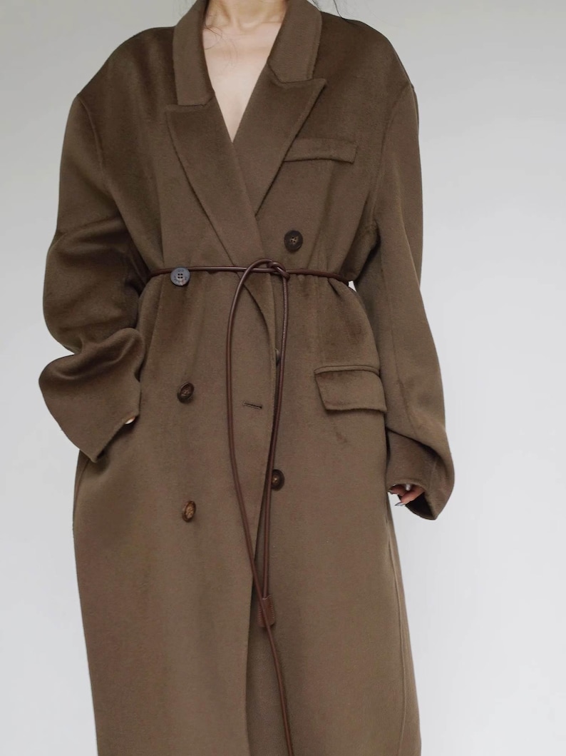 May include: An oversized olive brown wool-blend coat featuring a double-breasted front with dark brown buttons, wide lapels, and two side flap pockets. A thin, dark brown leather cord is tied around the waist as a belt, hanging down the front.