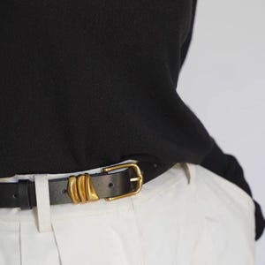 A person wears a black leather belt featuring a gold-tone buckle and three gold-tone decorative loops.