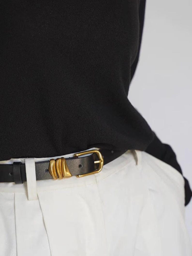 A person wears a black leather belt featuring a gold-tone buckle and three gold-tone decorative loops.