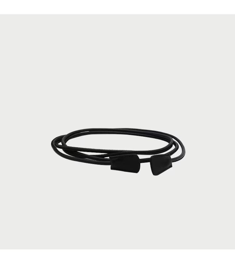 May include: A thin black cord belt featuring a smooth, rounded texture and two matching black, flat, tapered end pieces. The cord is coiled into multiple overlapping loops, creating a circular shape with the ends positioned side by side.