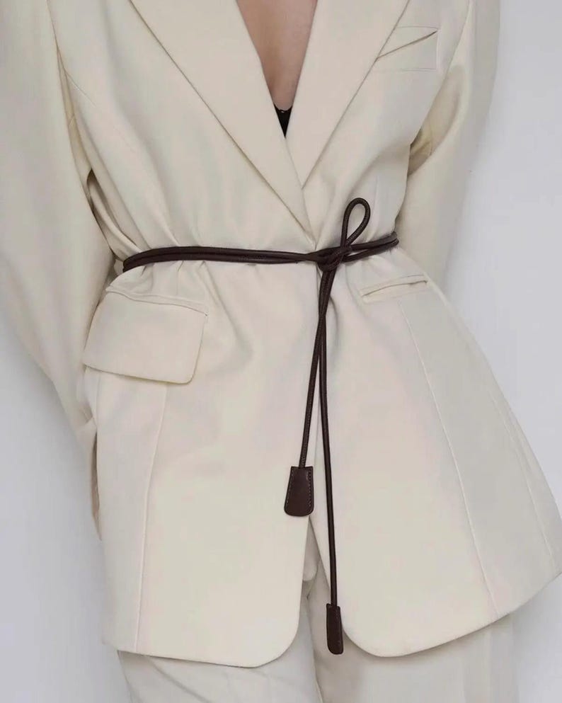 May include: A cream-colored blazer with notched lapels and two flap pockets, cinched at the waist with a thin, dark brown cord belt. The belt features two small, rectangular, dark brown end pieces that hang down from a simple knot at the front.