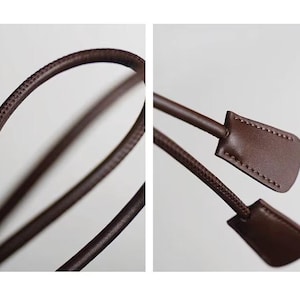 May include: A dark brown leather cord with a smooth, rounded profile and visible stitching along the edges. The cord features two flat, rectangular leather tabs at the ends, each secured with matching brown thread stitching.