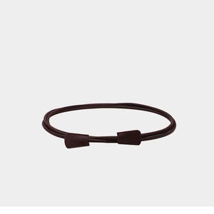 May include: A thin, dark burgundy belt featuring a smooth, rounded cord-like strap and two matching flat, trapezoidal-shaped end pieces that overlap at the front. The belt is arranged in a circular loop, showcasing a simple, minimalist design.
