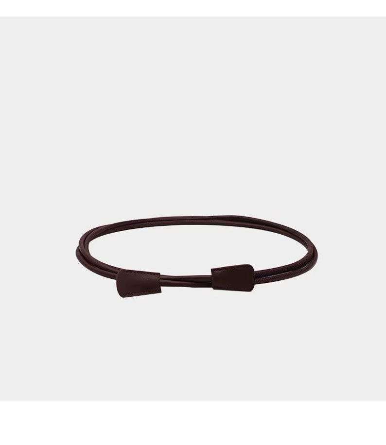May include: A thin, dark burgundy belt featuring a smooth, rounded cord-like strap and two matching flat, trapezoidal-shaped end pieces that overlap at the front. The belt is arranged in a circular loop, showcasing a simple, minimalist design.