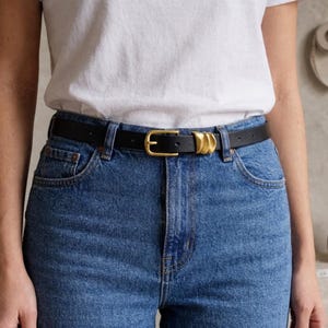 A person wears a black leather belt featuring a gold-tone buckle and three gold-tone decorative loops.