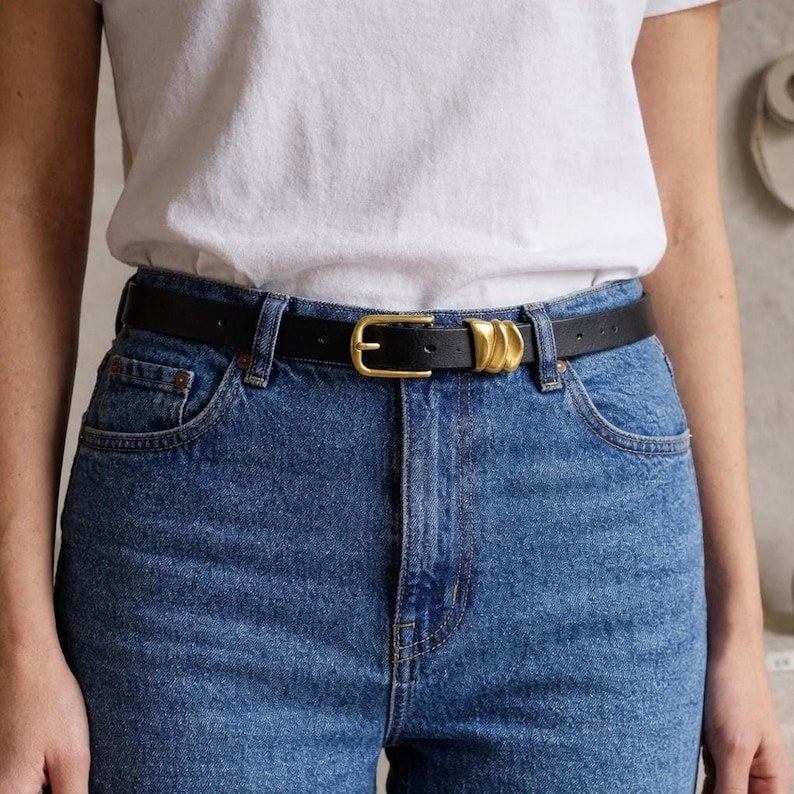 A person wears a black leather belt featuring a gold-tone buckle and three gold-tone decorative loops.