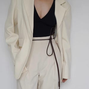 May include: A cream-colored blazer worn over a black V-neck top and cream-colored trousers. A thin, dark brown cord belt is tied around the waist of the trousers, featuring a long, dangling end with a small, cylindrical brown weight at the tip.