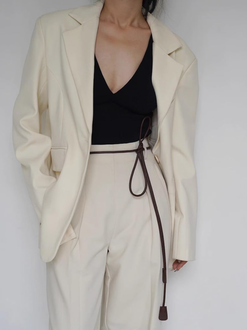 May include: A cream-colored blazer worn over a black V-neck top and cream-colored trousers. A thin, dark brown cord belt is tied around the waist of the trousers, featuring a long, dangling end with a small, cylindrical brown weight at the tip.