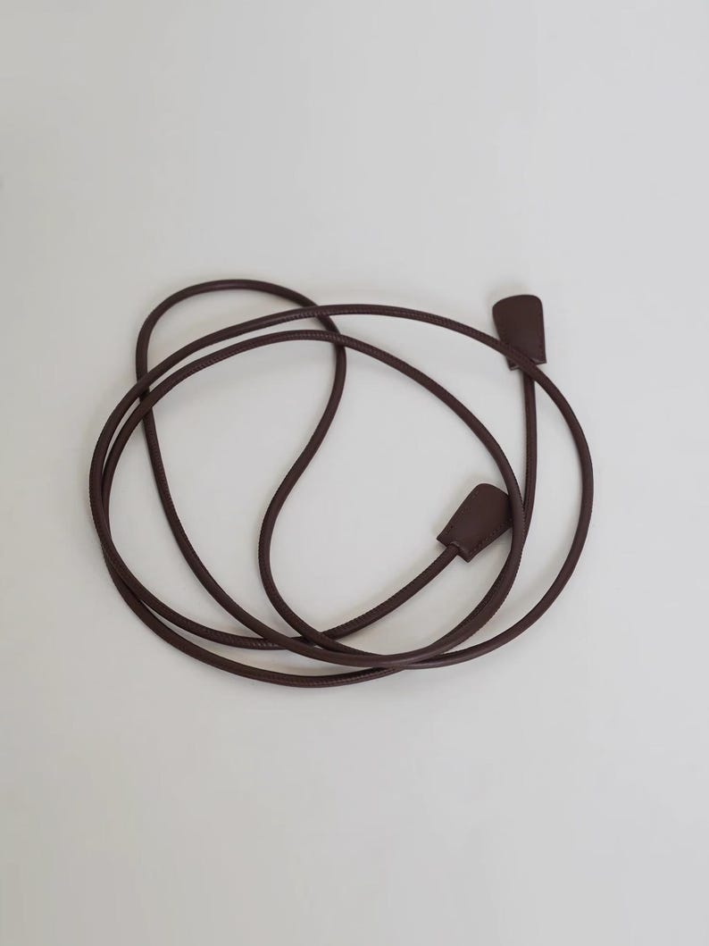 May include: A long, thin, dark brown cord strap with a smooth, rounded texture. The strap features two flat, rectangular-shaped end pieces in a matching dark brown color, securely attached to each end of the flexible, coiled cord.
