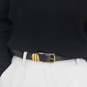 A person wears a black leather belt featuring a gold-tone buckle and three gold-tone decorative loops.