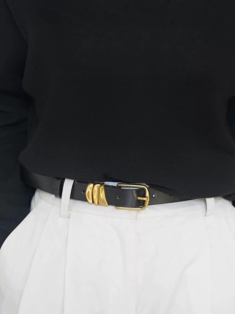 A person wears a black leather belt featuring a gold-tone buckle and three gold-tone decorative loops.