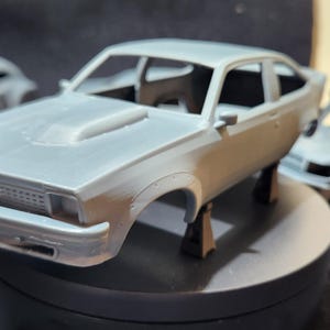May include: A collection of four unpainted model cars, including a station wagon, a saloon, a coupe, and a pick-up truck. The cars are made of grey plastic and are in various stages of assembly.