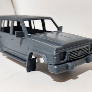 以下が含まれることがあります： グレーのプラスチック製スポーツユーティリティビークル（SUV）のモデル。モデルは、詳細なフロントグリル、サイドミラー、ルーフラックを備えています。ホイールは取り付けられていません。車両は明るいグレー色です。