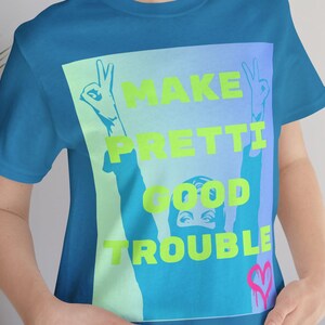 Può includere: T-shirt turchese con la scritta "MAKE PRETTI GOOD TROUBLE" in verde neon. Il disegno presenta una persona con le braccia alzate nel segno della pace. Un cuore rosa si trova nell'angolo in basso a destra.