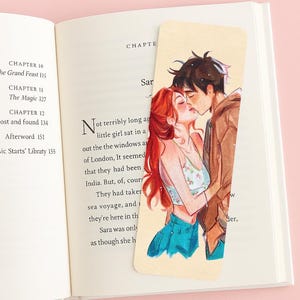 May include: A bookmark featuring an illustration of a couple kissing. The woman has long red hair, and the man has dark hair. The bookmark is placed in an open book with text visible.