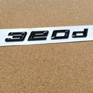 Op de afbeelding: Glanzend zwart "320d" embleem op een witte strip. De letters zijn in een modern schreefloos lettertype, met een subtiel 3D-effect. Waarschijnlijk een automodel.