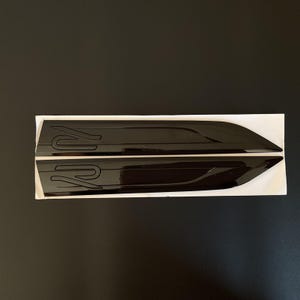 Pode incluir: Duas peças de acabamento automotivo pretas brilhantes com um design estilizado "R". As peças estão sobre um suporte branco, contra um fundo escuro. O acabamento tem uma forma elegante e aerodinâmica, sugerindo uma estética esportiva.